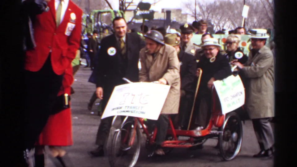 Older participants in a Wisconsin parade ride a red tricycle-style vehicle with hand-lettered signs while spectators and other marchers stand nearby.