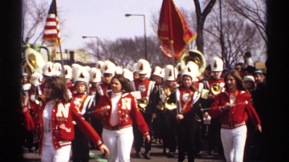 A red-and-white marching band in white helmets approaches during the 1971 Wisconsin parade, with flags, brass instruments, and spectators visible.