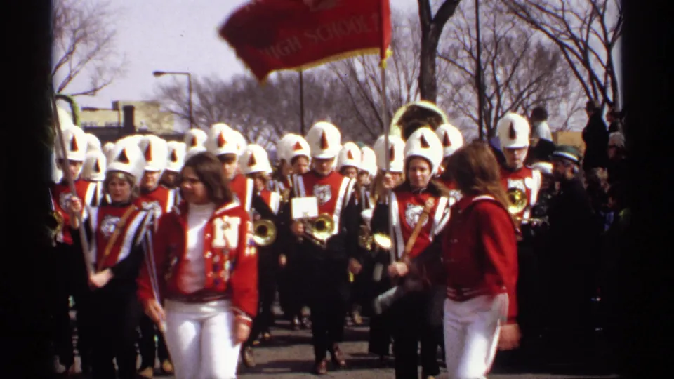 Band members in red uniforms and white helmets continue through the Wisconsin parade, partially framed by dark film-edge occlusion and bare trees.