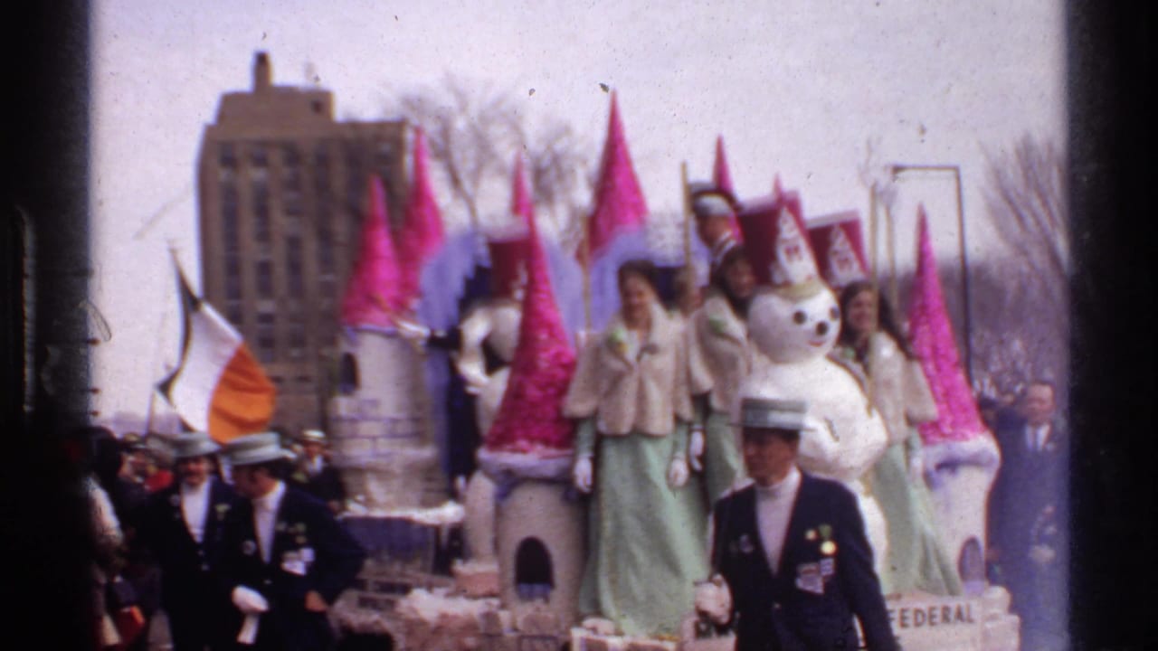 Snowman float and castle turrets pass by with riders, marchers, flags, spectators, and urban buildings behind.