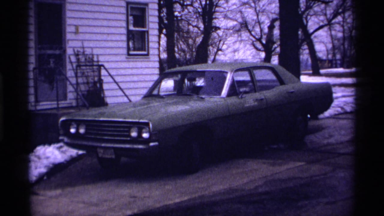 Greenish 1970s sedan parked in a driveway beside a white-sided house with snow nearby.