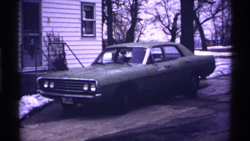 Greenish sedan in a Wisconsin residential driveway, with a white-sided house, bare trees, and remaining snow visible in the 1971 home movie frame.