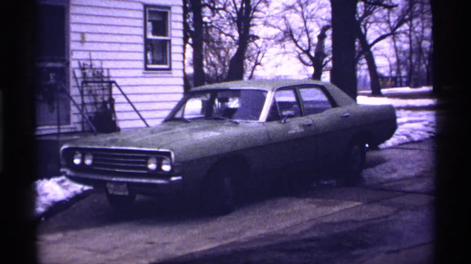 Late-winter 1971 Wisconsin frame showing a parked greenish sedan by a white-sided house, with snow along the driveway and leafless trees beyond.