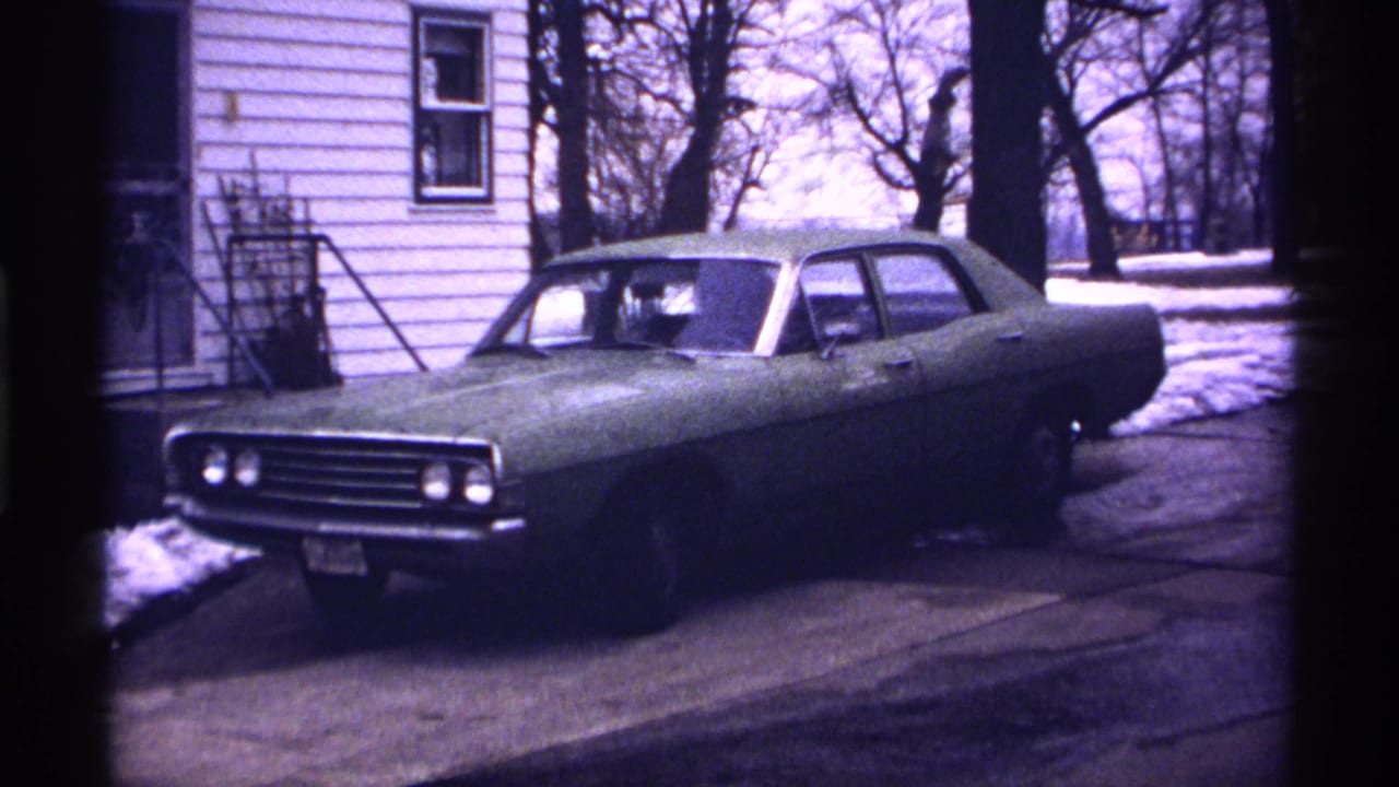 Classic sedan parked on a snowy driveway beside a white-sided house.
