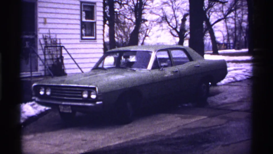 1971 Wisconsin home-movie frame showing a classic American sedan on a snowy residential driveway near a white-sided house.