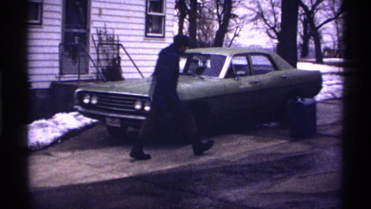 Man in a dark coat walks toward a green car parked by a white-sided house.