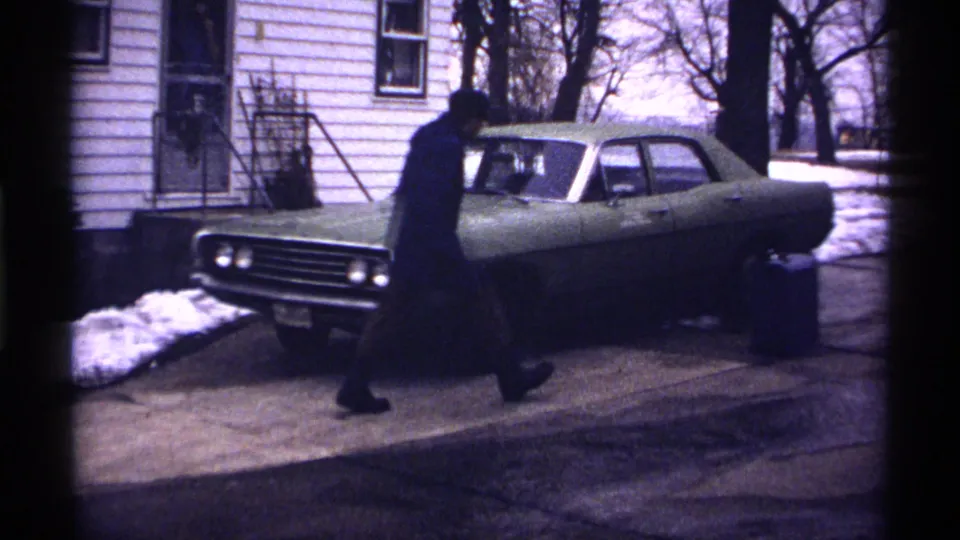 In 1971 Wisconsin home-movie footage, a man in a dark coat approaches a green car beside a white-sided house with snow nearby.