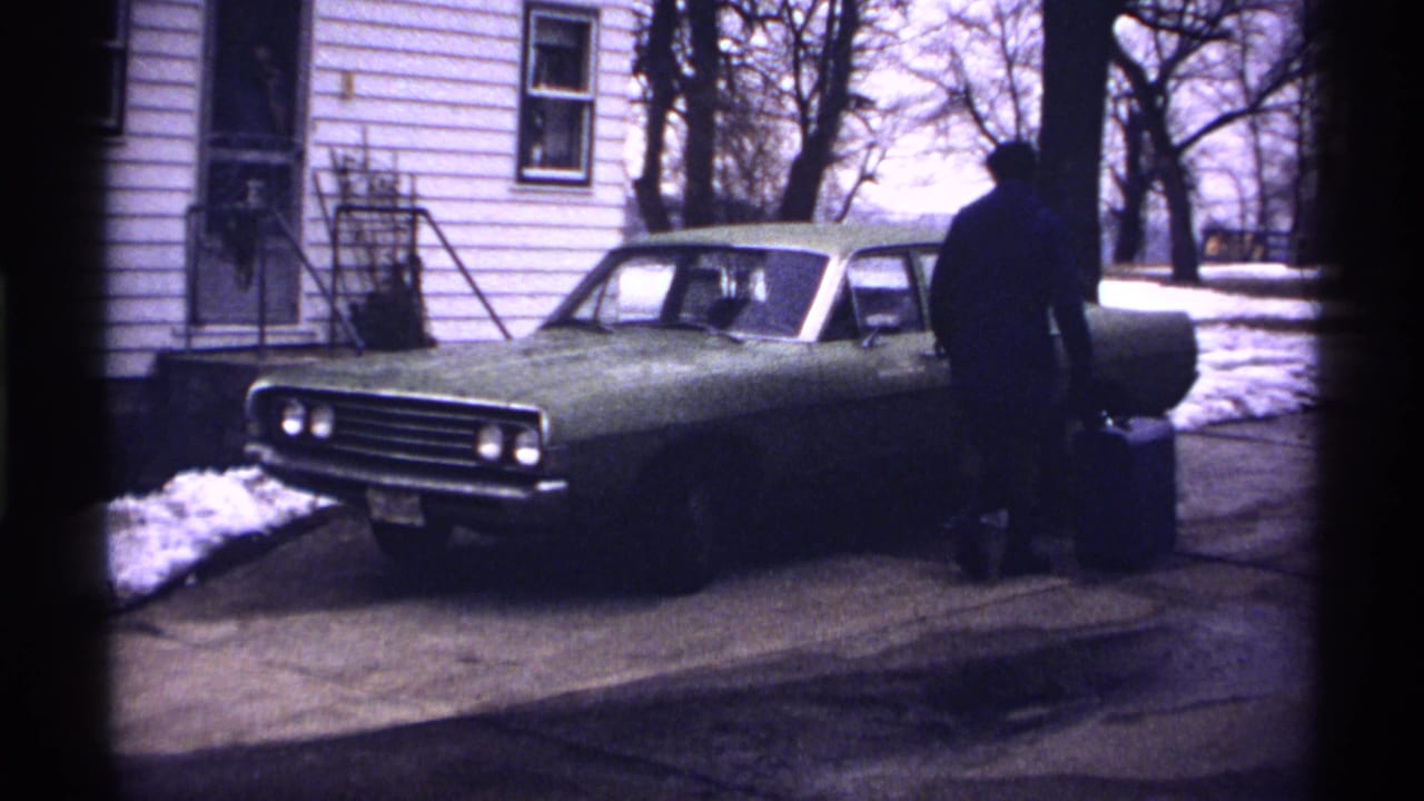 Man stands near the driver's side of a green car outside a house in winter.