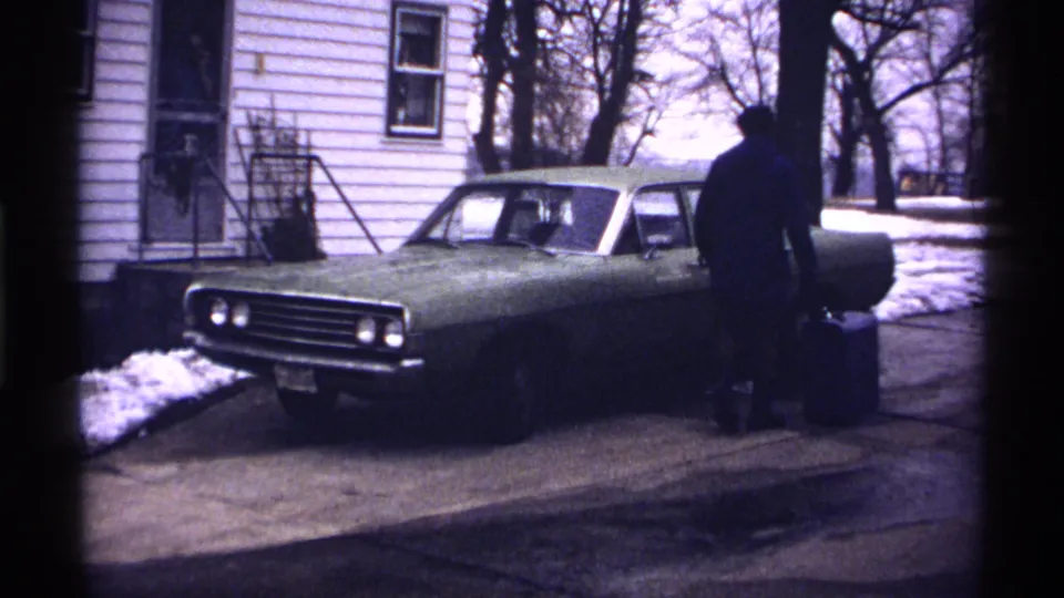 The man reaches the driver's side area of the green car, with the white house, bare trees, and snow patches behind him.