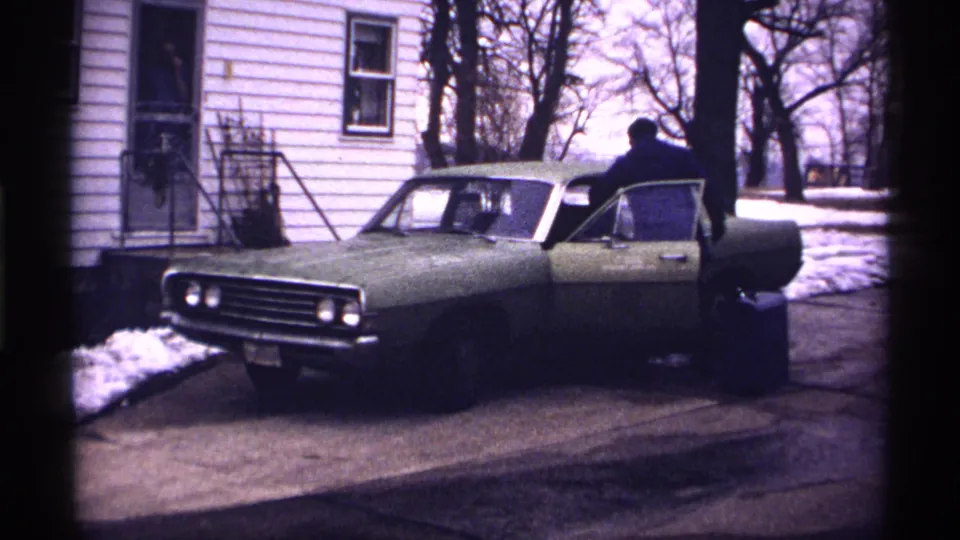 The man has opened the driver's door of the green car, with winter snow patches and the white-sided house visible around him.