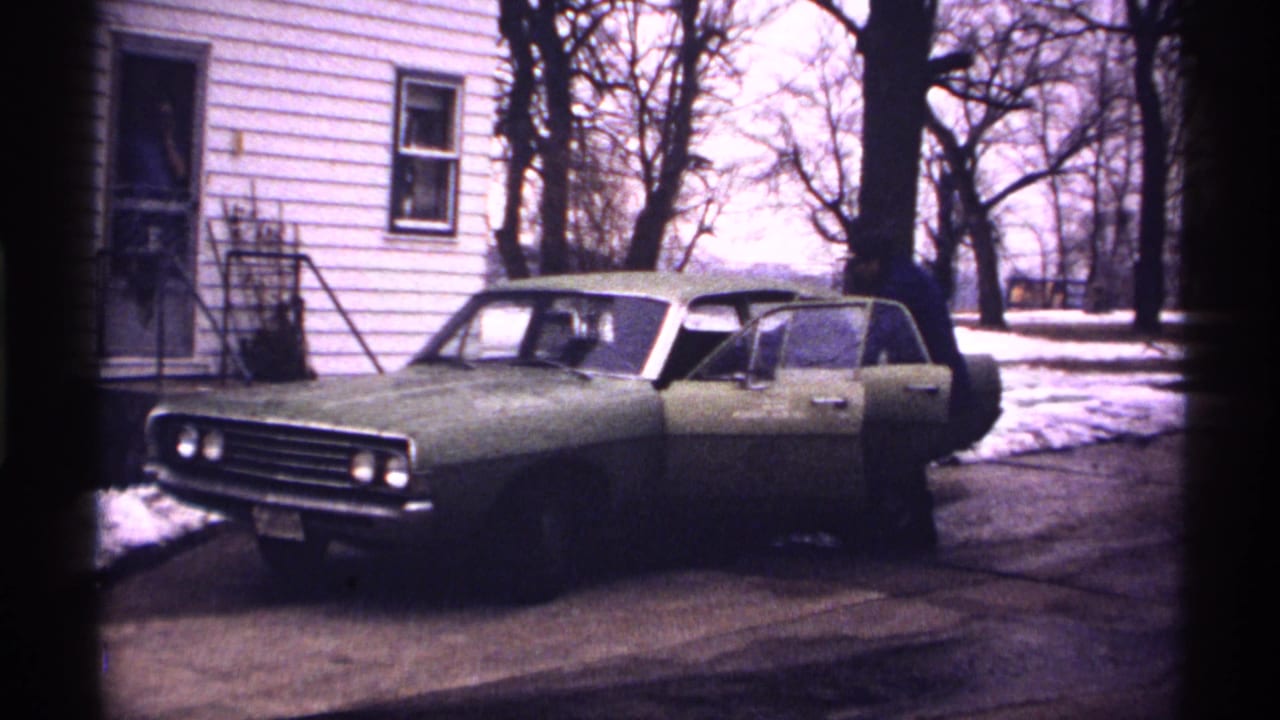 Green sedan in a snowy driveway with a man at the open driver's door.
