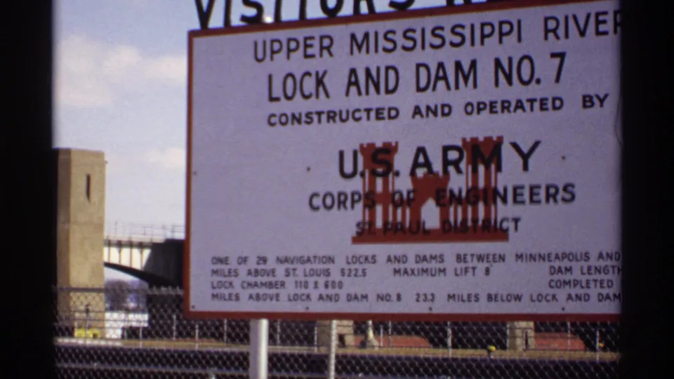 Close 8mm view of the Upper Mississippi River Lock and Dam No. 7 sign, operated by the U.S. Army Corps of Engineers, with fencing and structures behind.