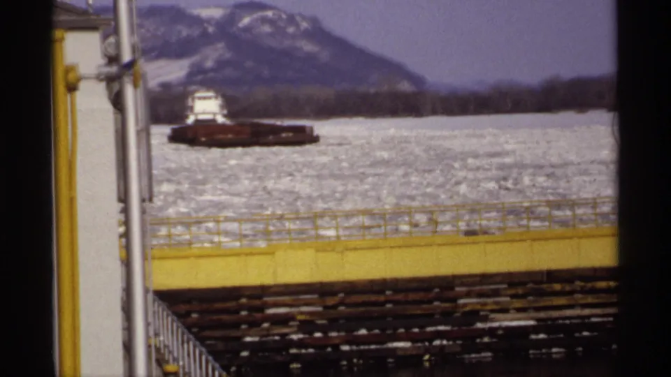 Wide 1971 home-movie view of a barge on icy Upper Mississippi River water, with a distant bluff or hill and lock structure in the foreground.