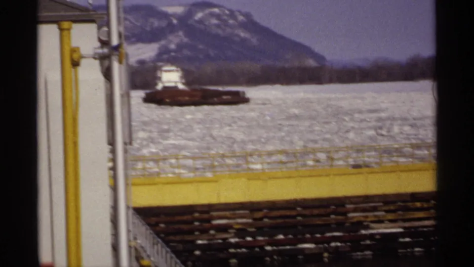 A soft, grainy 8mm view shows the barge on slushy winter water, partially framed by a yellow waterfront railing and pipes.