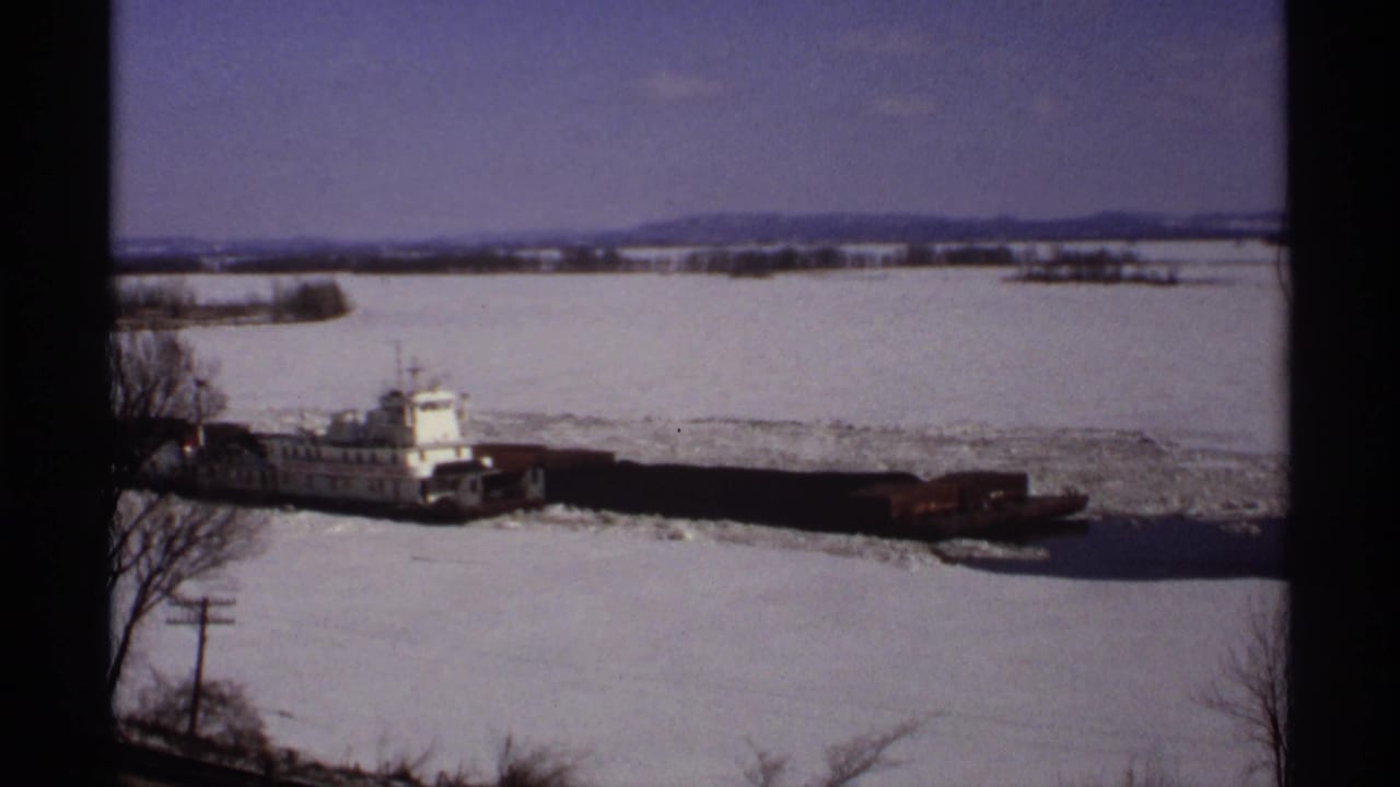 1971 Wisconsin 8mm frame showing a white tugboat and dark barges moving through a partially frozen river with snow-covered banks.