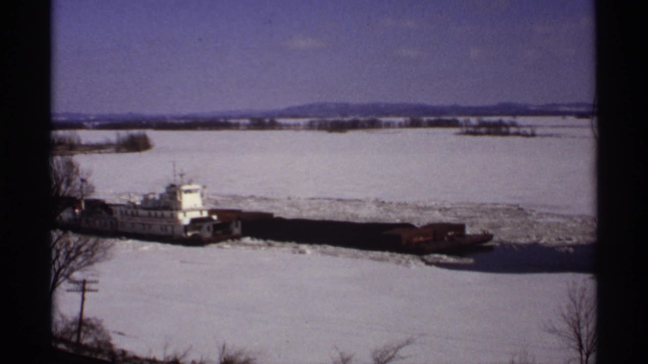 Tugboat and barges continue across a frozen river bordered by snow.