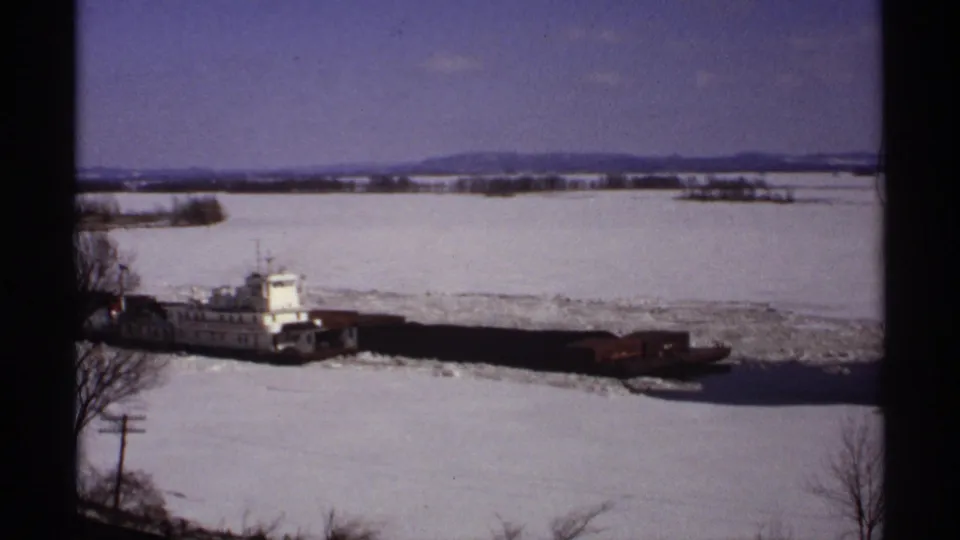 The tug and barge line passes through icy water in a Wisconsin winter scene, with distant hills and bare shoreline trees visible.