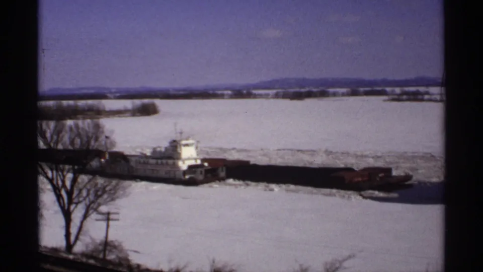 1971 home-movie view of a tugboat pushing barges through river ice, with bare trees, utility poles, snow, and low hills in the background.