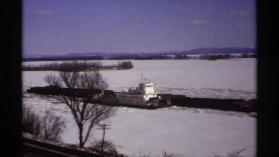 A Wisconsin winter river view shows a tug and barges partly framed by bare branches as they move through broken ice.