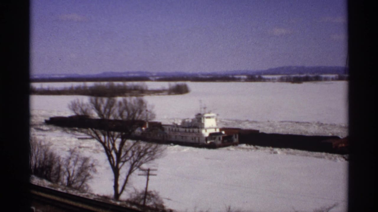 1971 Wisconsin 8mm frame of a white tugboat pushing barges through ice on a snow-covered river, viewed through dark window framing.