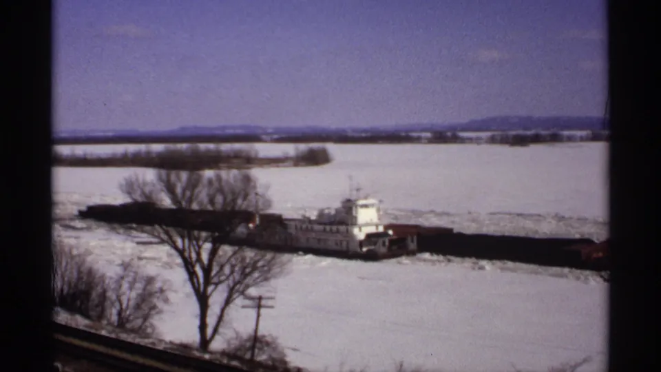 1971 Wisconsin 8mm frame of a white tugboat pushing barges through ice on a snow-covered river, viewed through dark window framing.