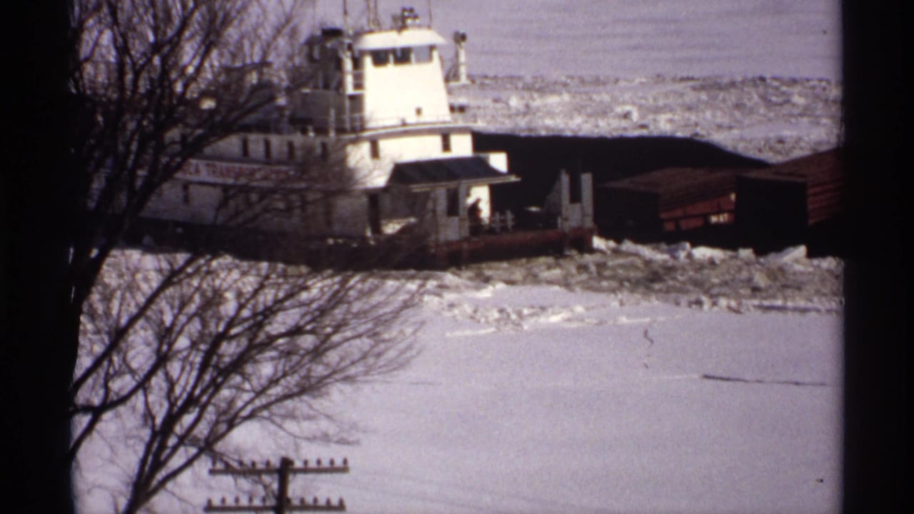White tugboat and barges pass behind bare trees on icy water.