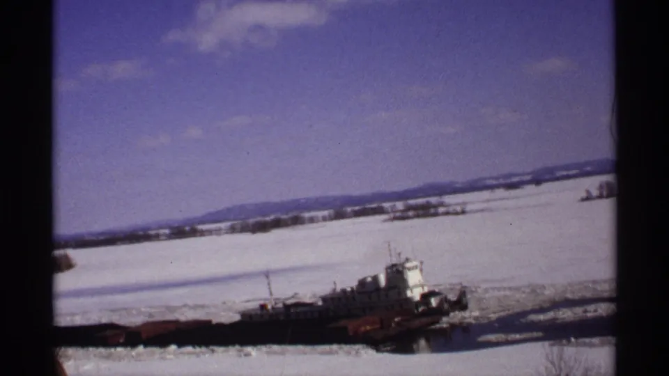 A wider 1971 Wisconsin frame shows the tug and barges on an icy river below pale sky and distant hills, still partly framed by the window.