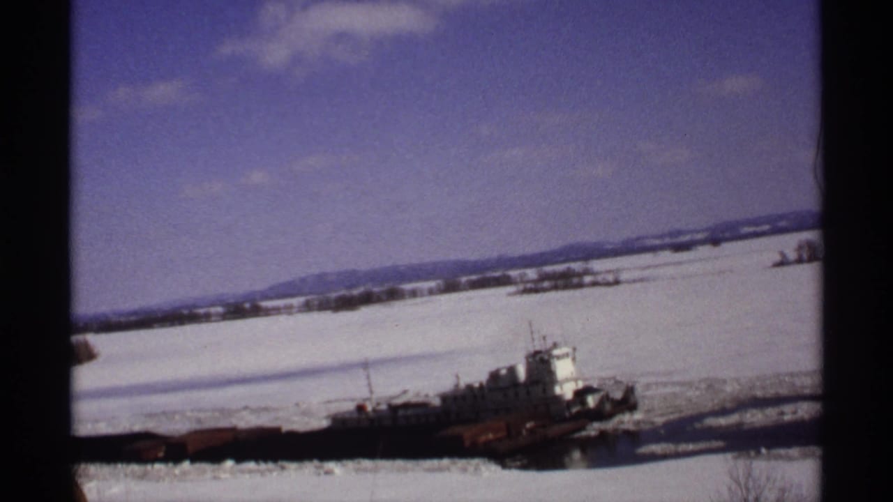 Tugboat and barge moving through ice on a snowy river under a blue sky.