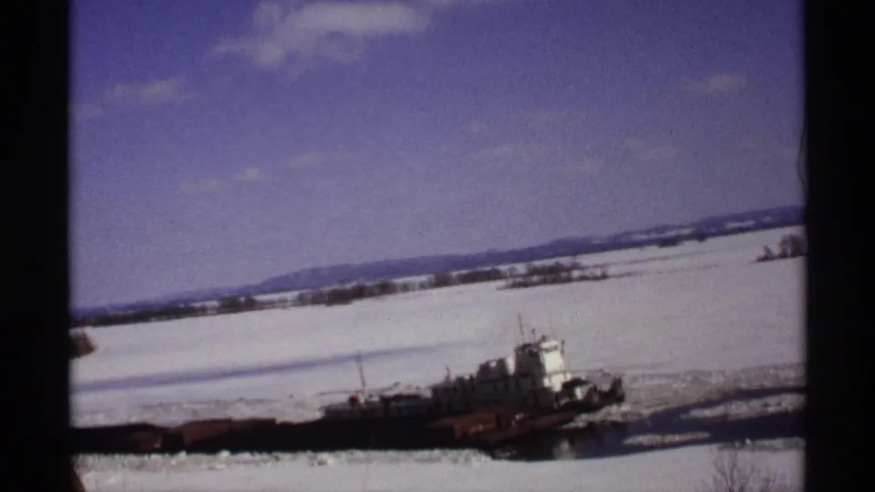1971 Wisconsin 8mm frame showing a tugboat towing a low barge through broken ice on a snow-covered river.