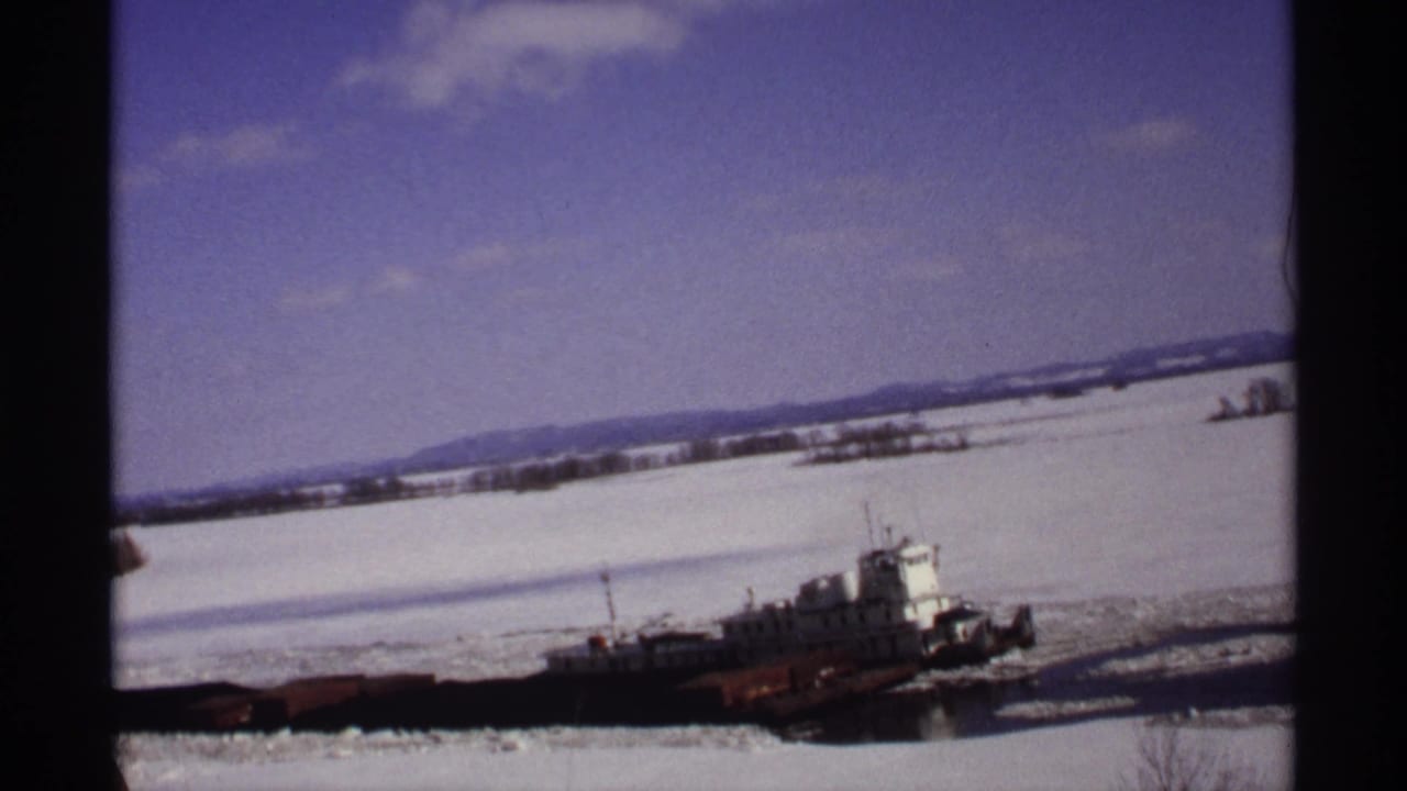 The tugboat appears farther along the snowy Wisconsin river, cutting through ice beneath scattered clouds.