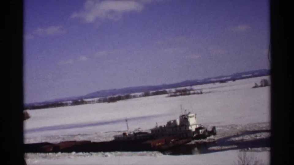 The tugboat appears farther along the snowy Wisconsin river, cutting through ice beneath scattered clouds.