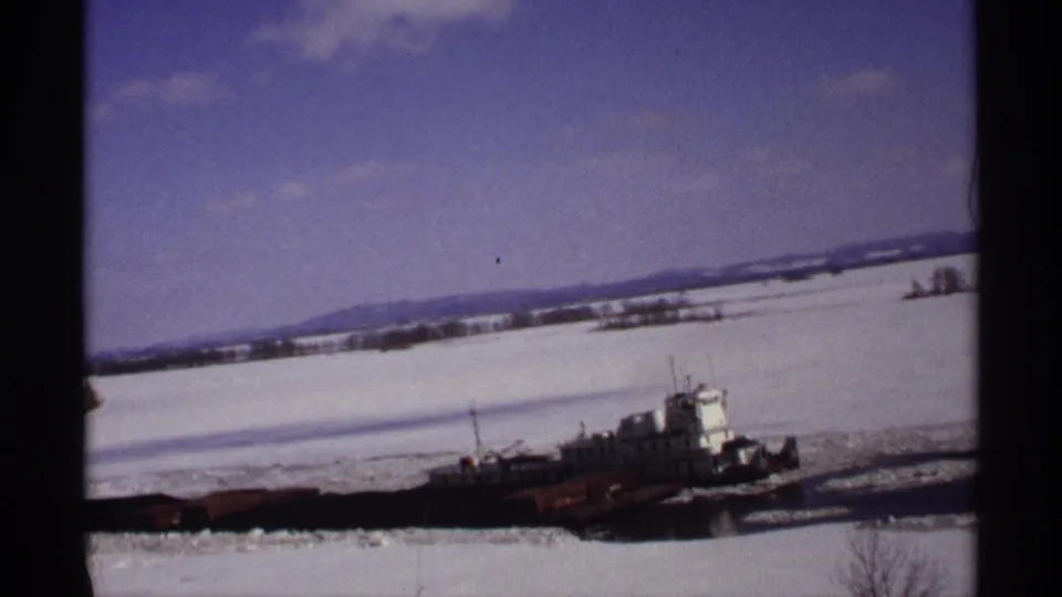 A home-movie view of the tugboat and barge crossing an icy Wisconsin river in bright winter daylight.
