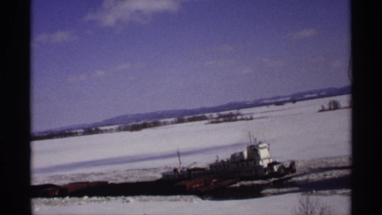 Tugboat moving through broken ice on a snowy river with distant hills and blue sky.