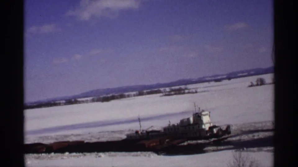1971 Wisconsin 8mm frame showing a tugboat crossing an icy river, with snow-covered banks, distant hills, and dark film borders.