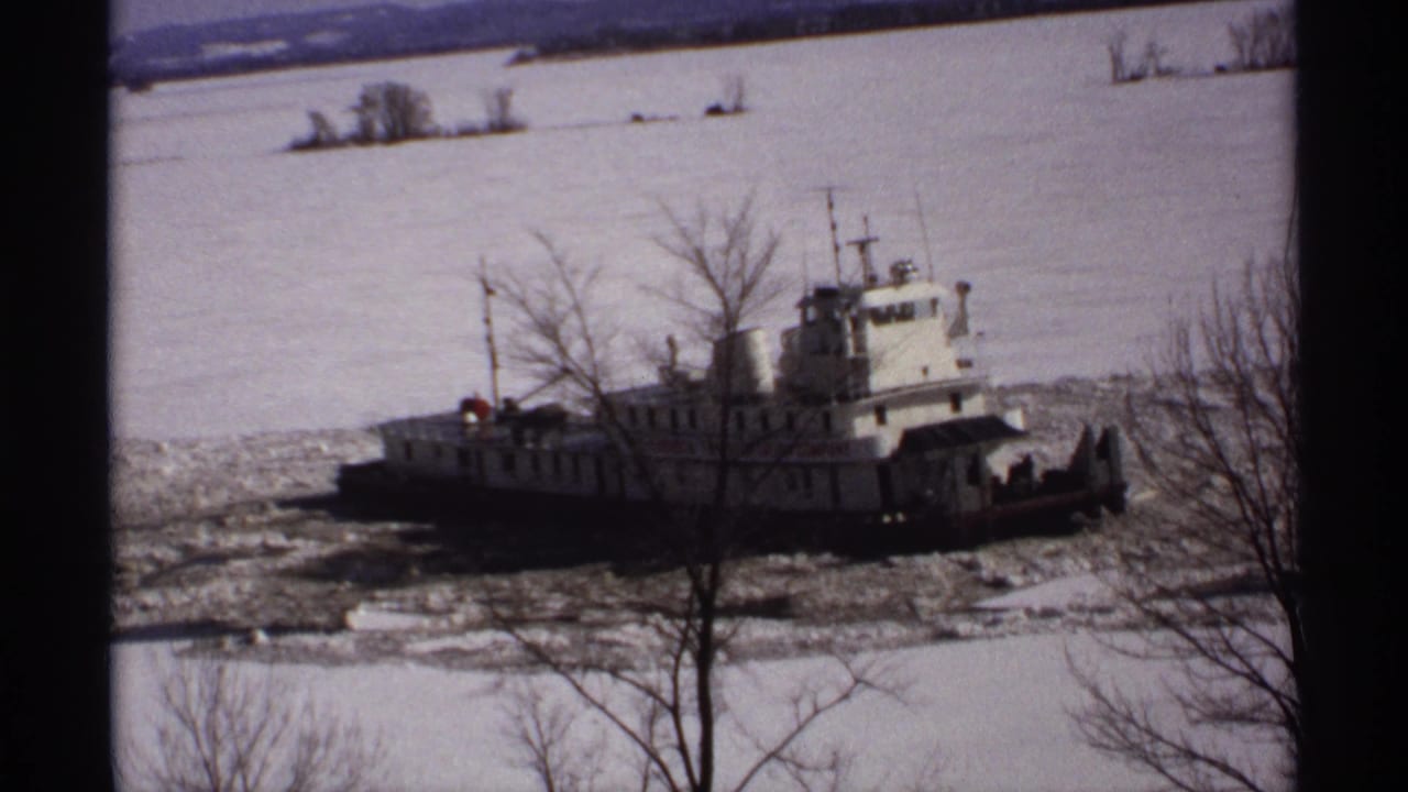 White tugboat near bare trees, surrounded by ice on a frozen river.