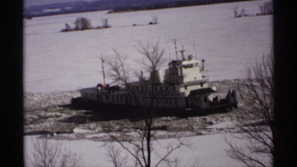 1971 Wisconsin 8mm frame with a white tugboat pushing through broken ice near leafless riverside trees and snowy banks.