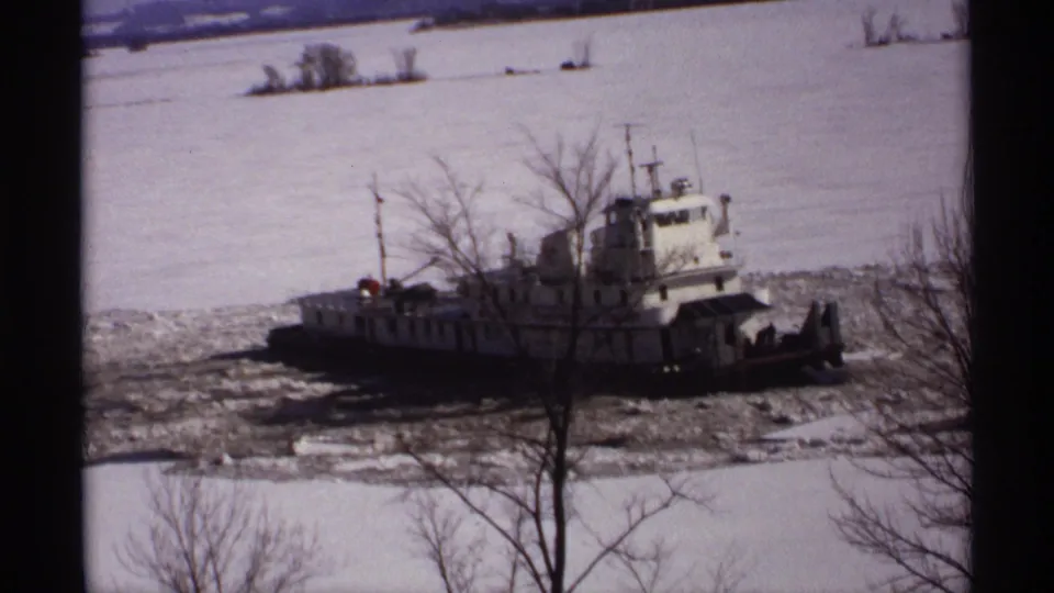 1971 Wisconsin 8mm frame showing a white tugboat in broken river ice, with snow, bare trees, and dark foreground bars.