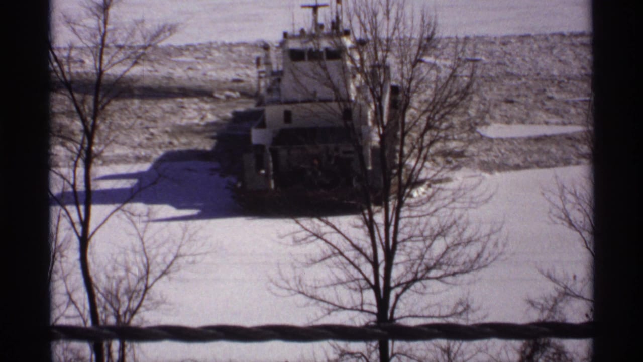 Tugboat bow faces the camera amid broken ice and snow, partly obscured by bare trees.