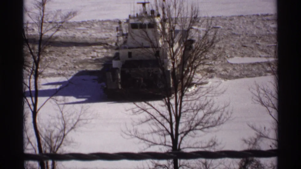 1971 Wisconsin winter home-movie frame showing the tugboat in broken ice behind tree branches and a dark foreground barrier.
