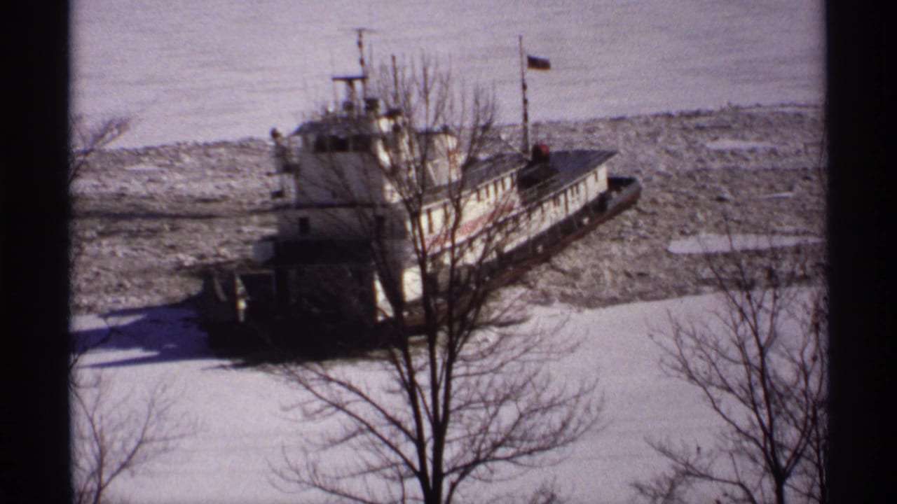 White tugboat moving through broken ice on a snowy river, partly blocked by tree branches.