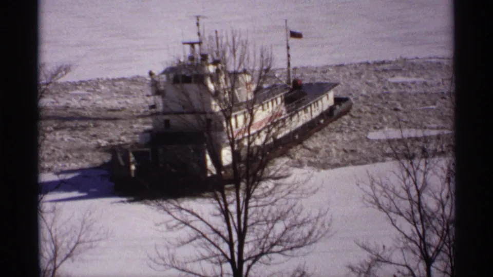 1971 Wisconsin home-movie frame of a white tugboat pushing through broken river ice under bright winter sun.