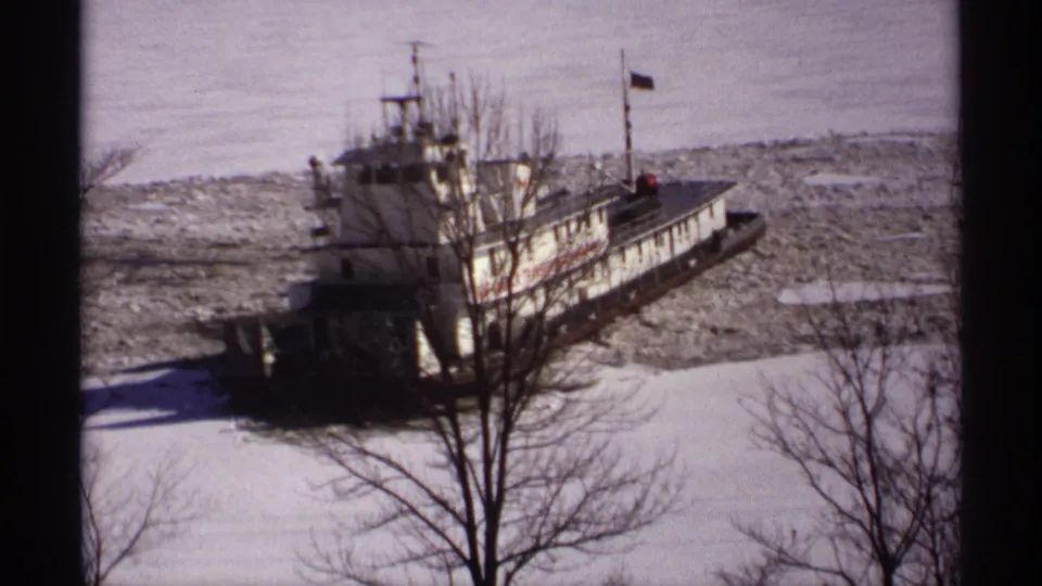 1971 Wisconsin 8mm frame with the tugboat more side-on, moving through broken ice behind leafless trees.