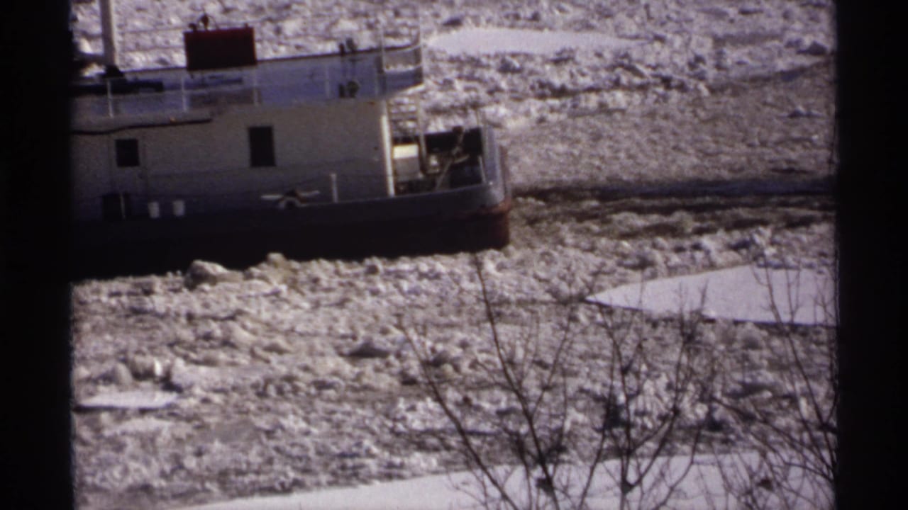 Tugboat partly framed by dark window edges while moving through broken river ice.