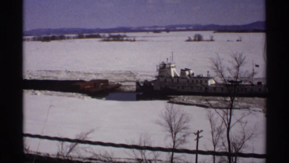 1971 Wisconsin 8mm frame with a wider snowy river view, a tugboat near shore, bare trees, distant land, and pale winter sky.