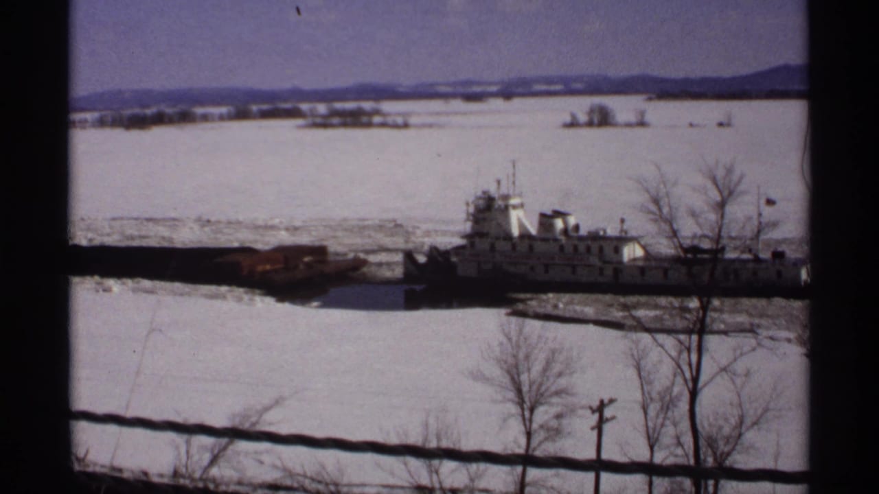 1971 Wisconsin 8mm frame of a white tugboat and barge passing through broken ice, seen past bare trees and dark window framing.