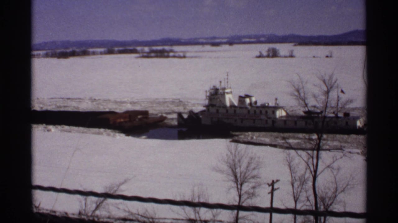 White tugboat beside a barge on icy water with bare trees in the foreground.