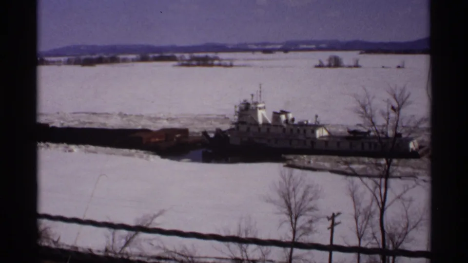 The tugboat and barge continue through broken ice, viewed from above with bare winter trees and distant hills in the background.