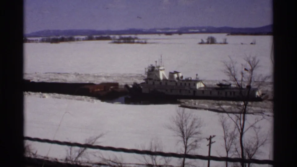 Final 1971 Wisconsin frame shows the white tugboat and barge partly obscured by bare trees as they pass through icy water.