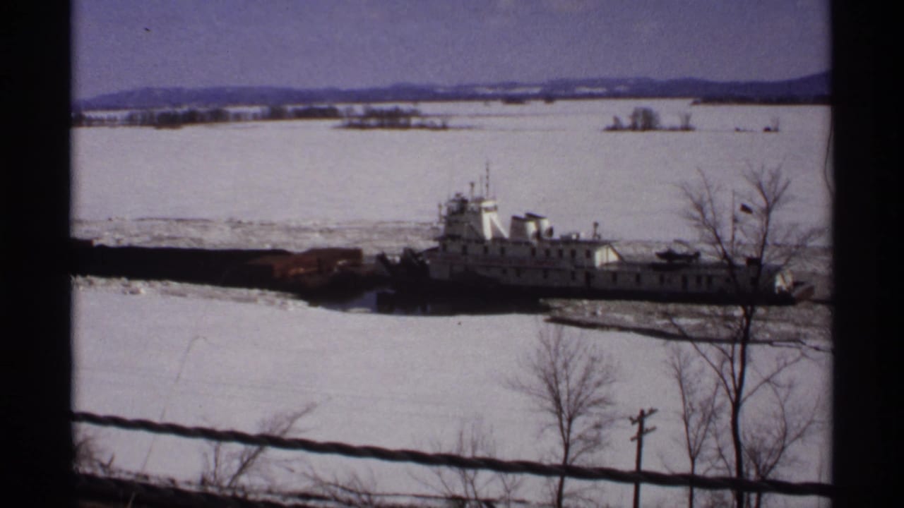 Icebreaker tug moves through frozen river ice behind a wire fence.