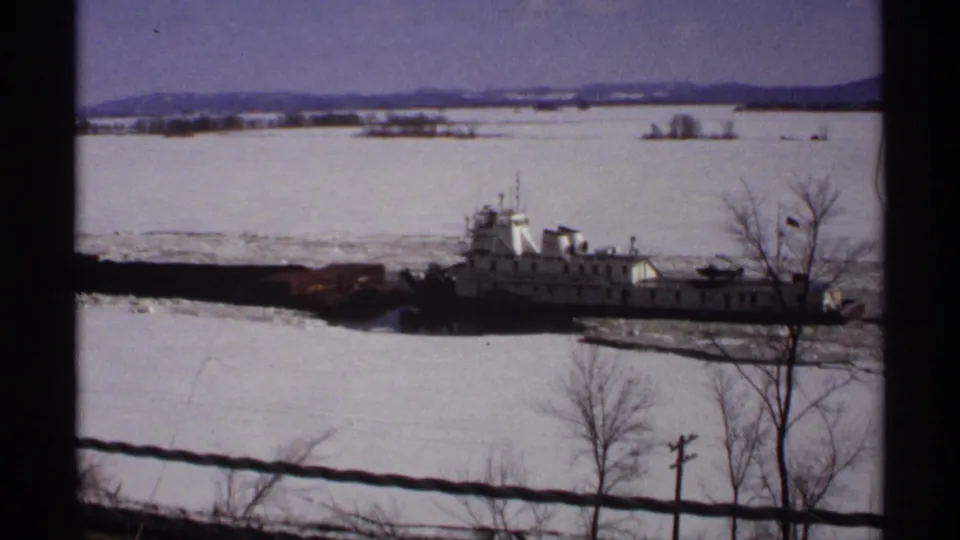 1971 Wisconsin 8mm frame showing a white and dark tugboat breaking ice on a frozen river, seen past a wire fence and bare trees.